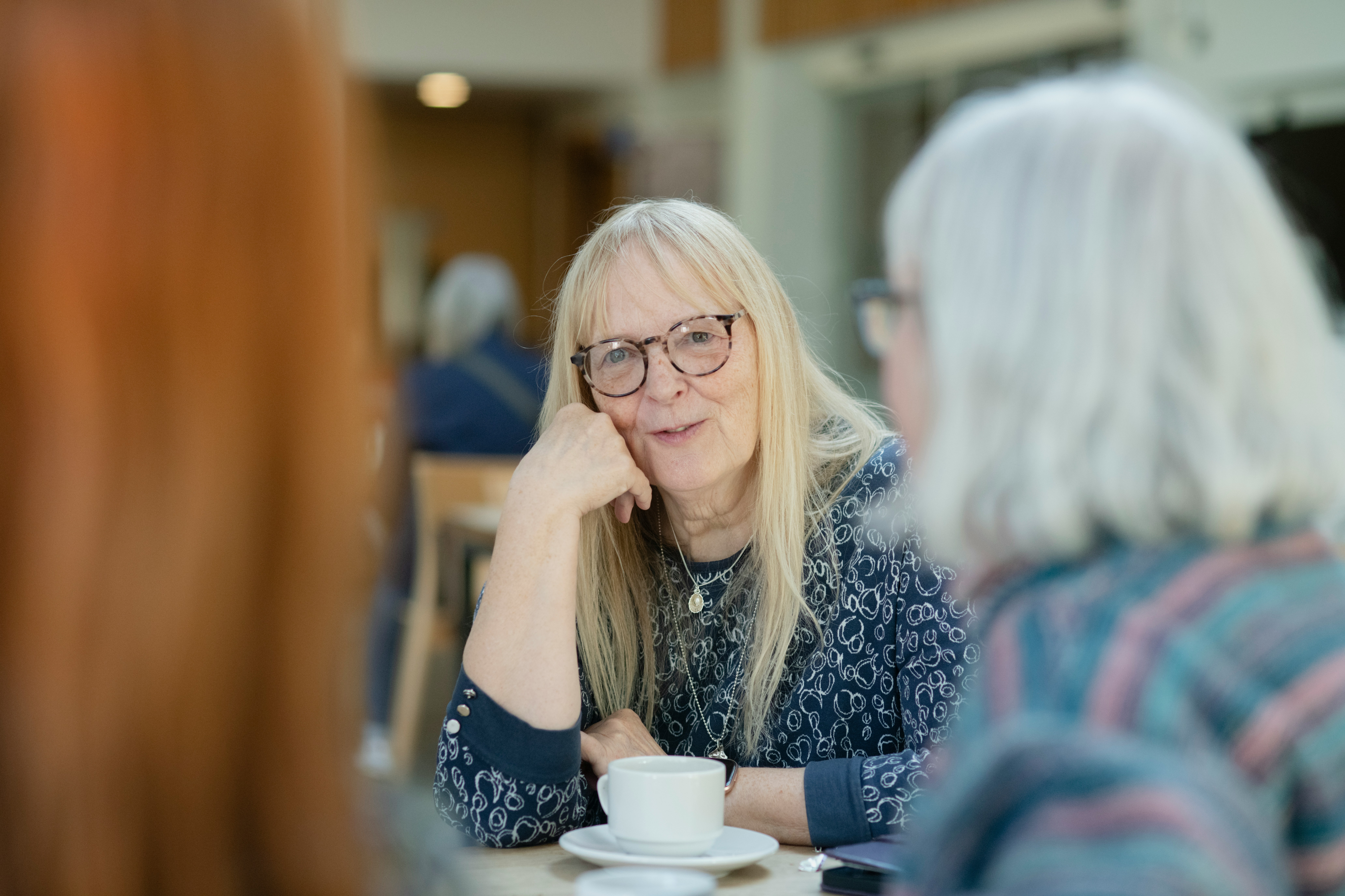 An older woman with glasses, with a cup of tea, mid conversation with another person who's back is to the camera